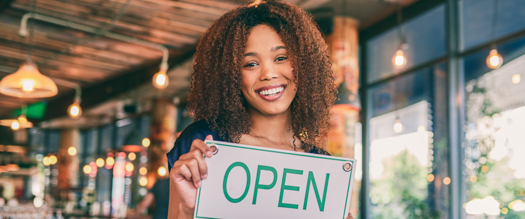 A young female business owner holds the open sign to her coffee shop.