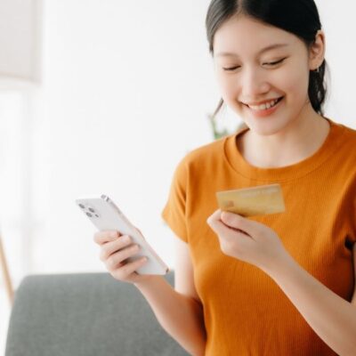 A young woman looking at her debit card while she's on her phone using her mobile banking app.